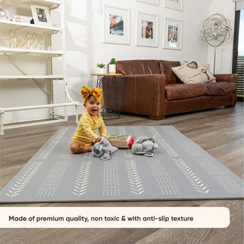 Baby sitting on a gray patterned mat in a living room with a brown sofa and toys.